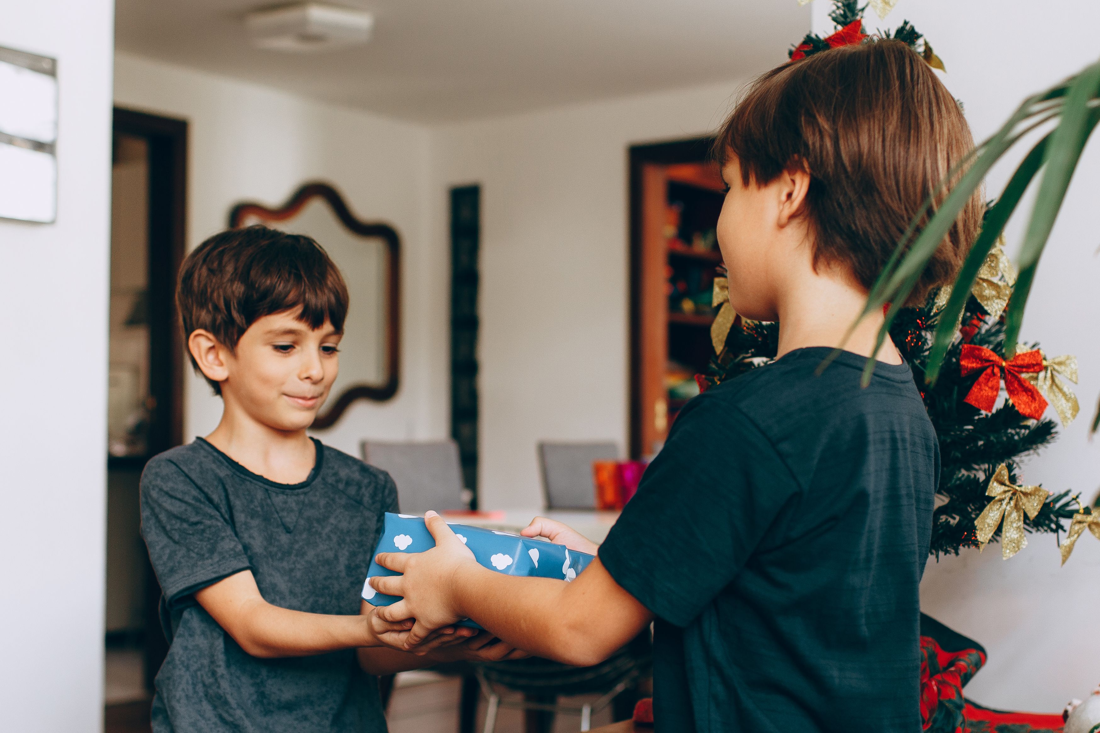 Two young boys exchanging gifts by a Christmas tree in a festive, indoor setting.