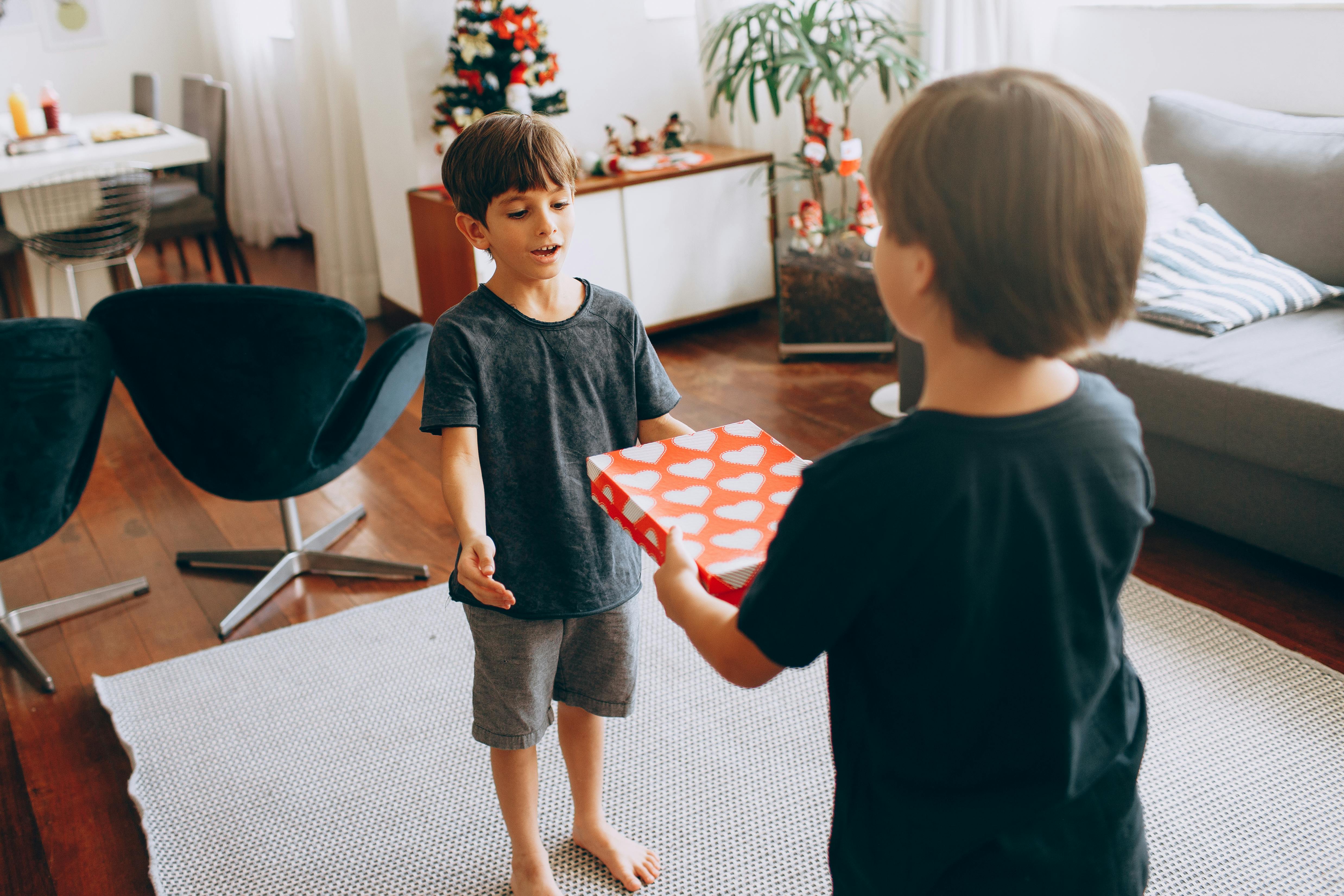 Two young boys exchanging a heart-patterned gift box in a cozy, festive living room setting.
