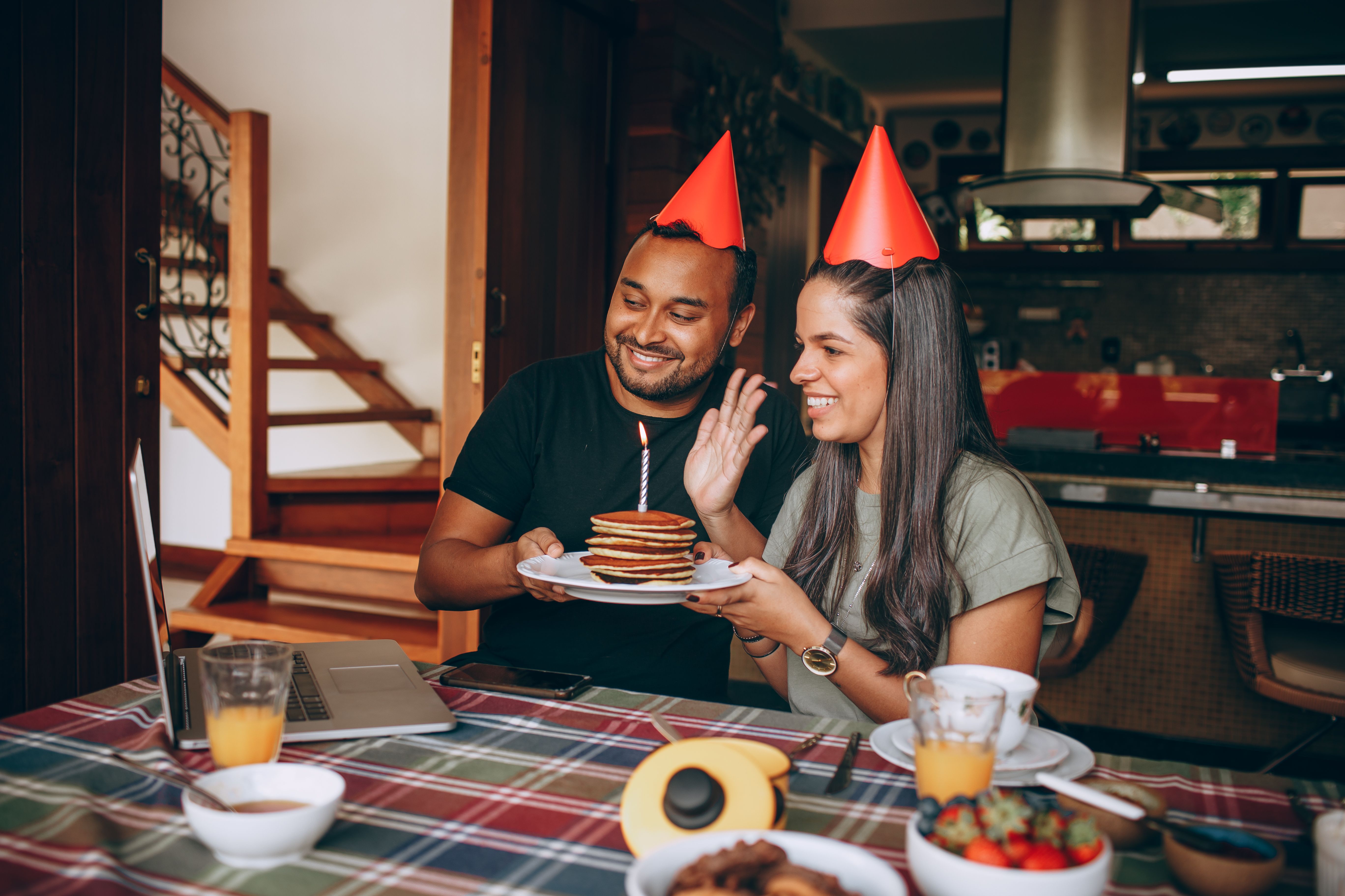 A joyful couple celebrating a birthday with pancakes and party hats indoors.