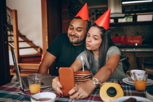 A couple celebrates a birthday at home with pancakes and party hats, capturing the moment with a selfie.