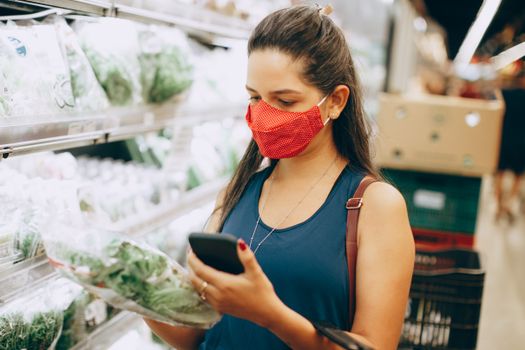 Woman wearing red face mask shopping for greens in a supermarket, checking her phone.