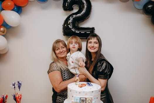 Three generations celebrating a child's second birthday with cake and balloons indoors.