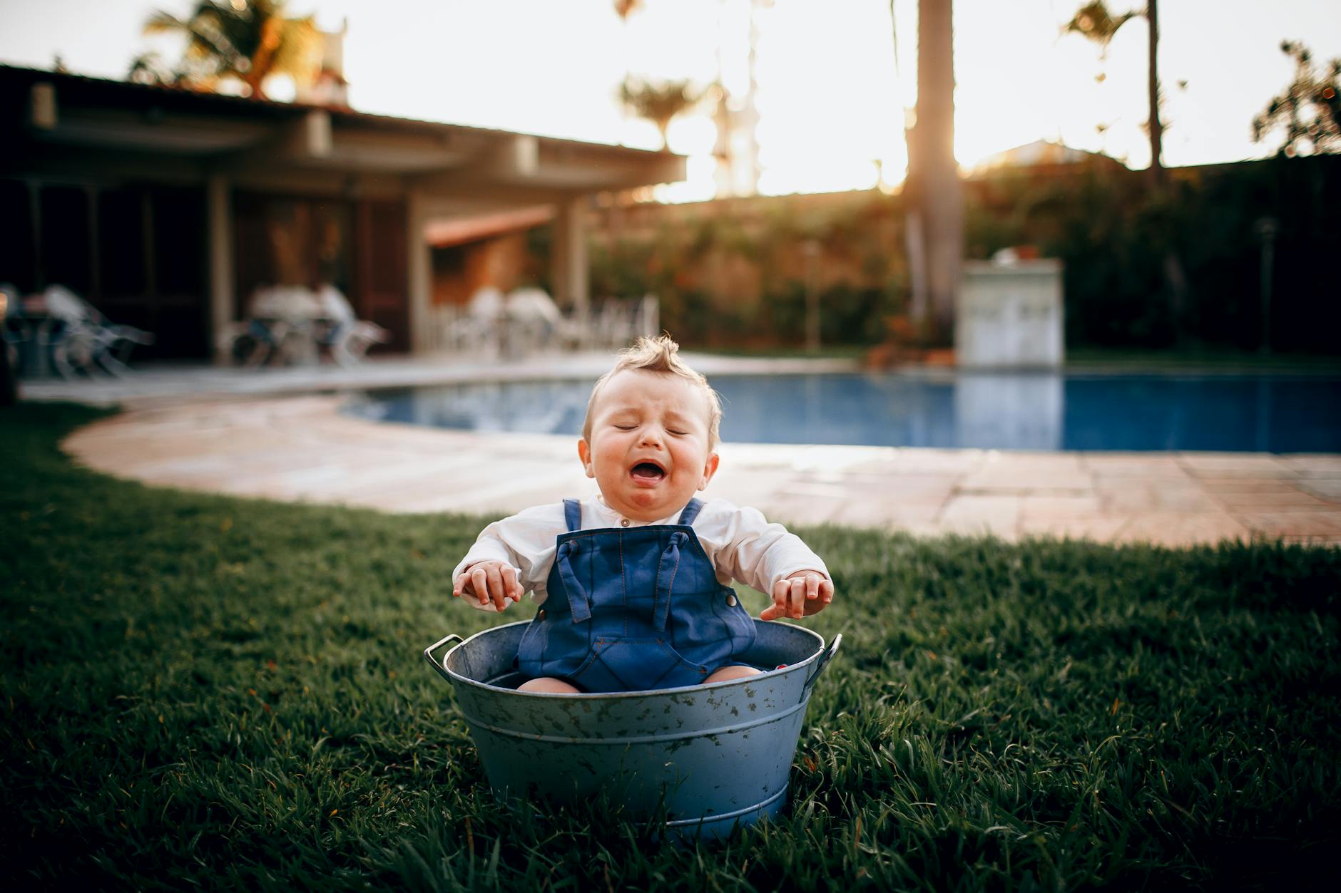 Adorable baby cries while sitting in a bucket by the pool during sunset.