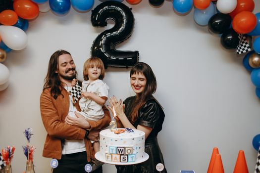 Family celebrating a child's second birthday with balloons and cake indoors.