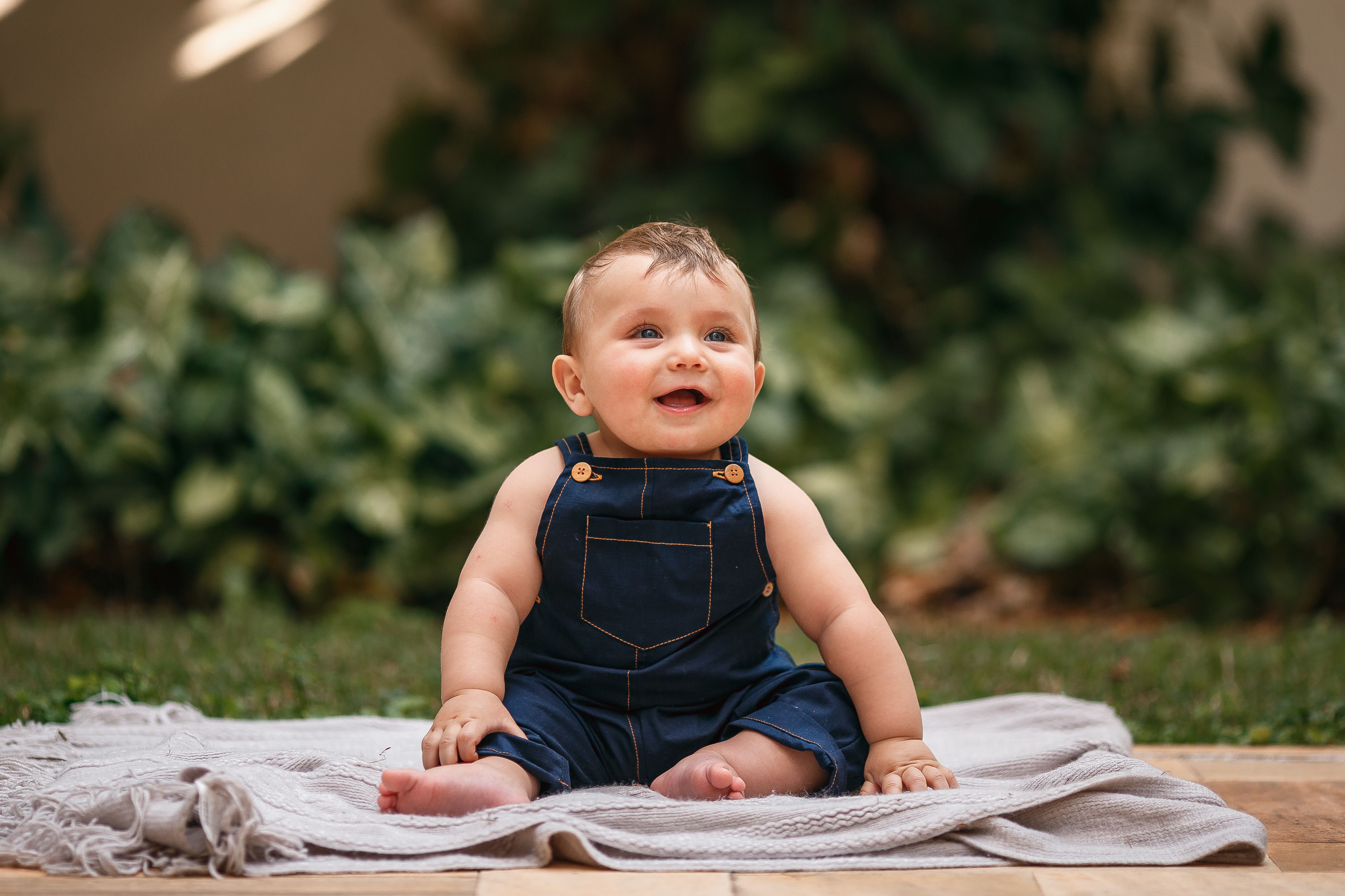 Adorable baby in overalls sitting on a blanket outdoors, smiling brightly.