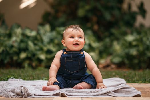 Adorable baby in overalls sitting on a blanket outdoors, smiling brightly.