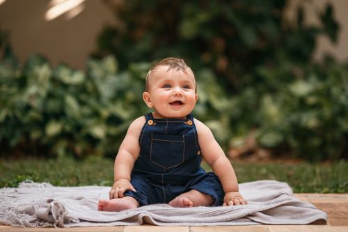 Free Adorable baby in overalls sitting on a blanket outdoors, smiling brightly. Stock Photo