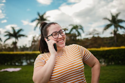Smiling woman in striped shirt talking on phone outdoors with palm trees in the background.
