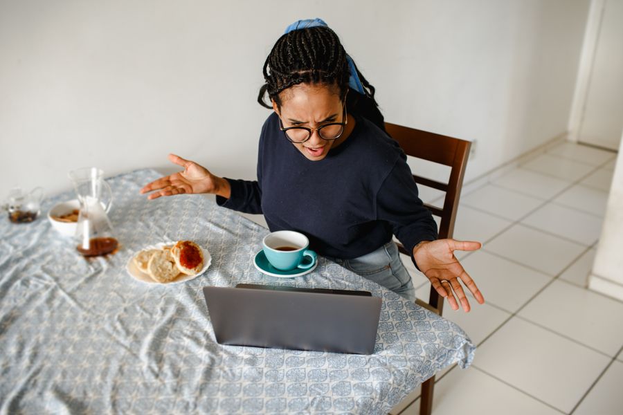 Woman gesturing in frustration while working on a laptop at home with coffee and breakfast.