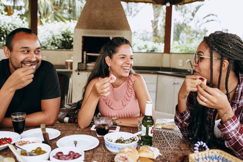 Free Three friends laughing and enjoying a meal outdoors with drinks and food on the table. Stock Photo