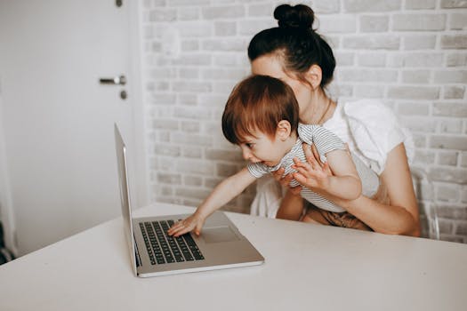 A young mother holds her baby while they both engage with a laptop, depicting modern parenting.