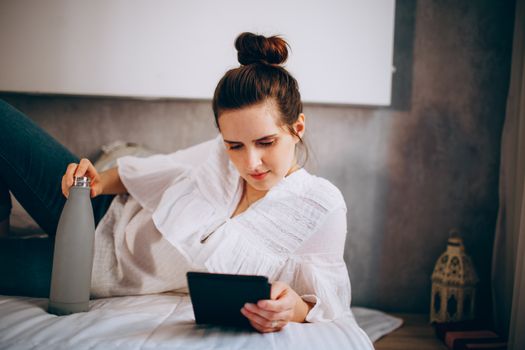 A woman in casual wear reads a tablet while relaxing indoors on a bed with a water bottle.