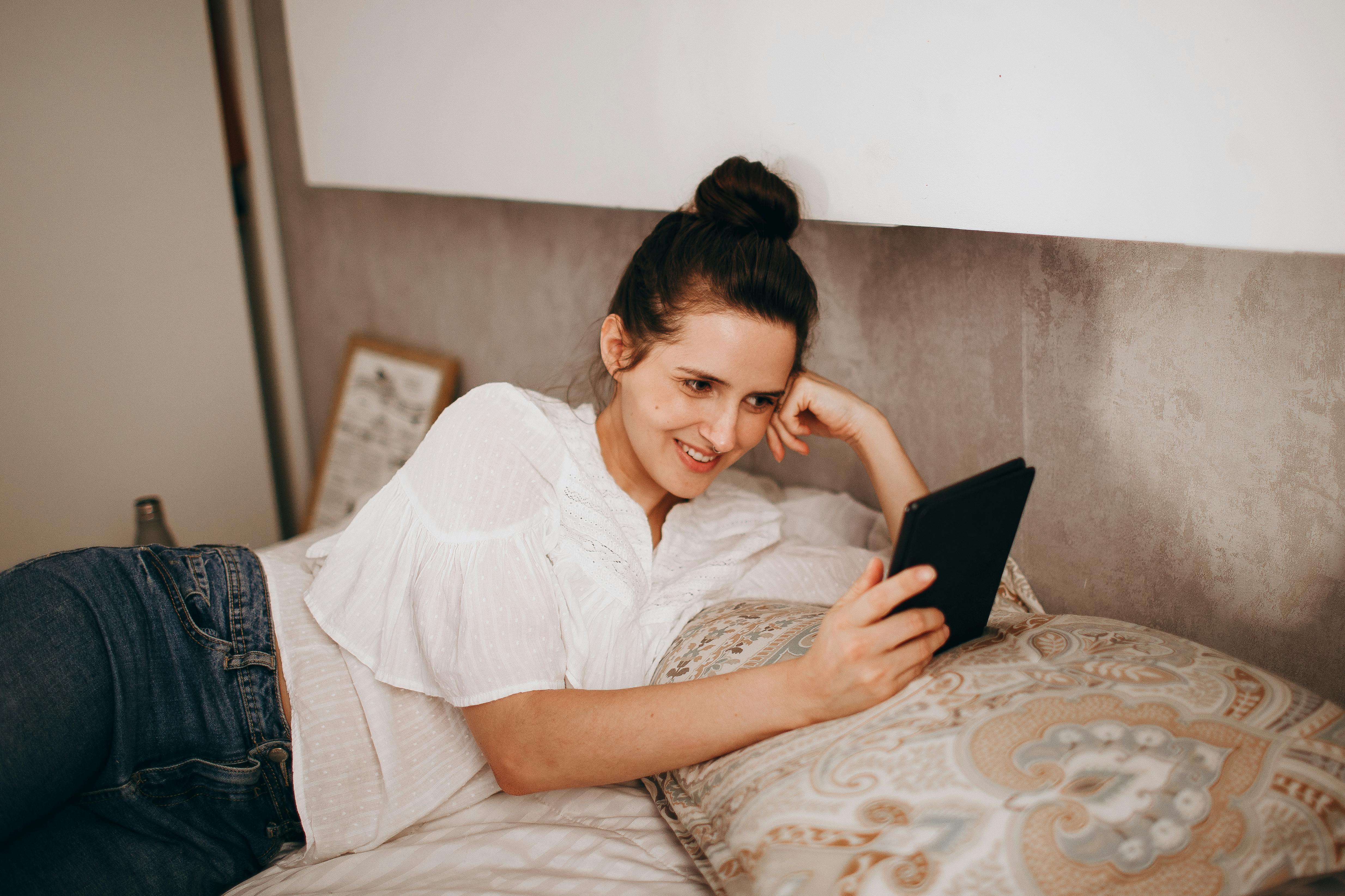 Adult woman relaxing on bed with an e-reader, enjoying leisure time in a cozy bedroom setting.