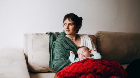 A mother sitting on a sofa holding her sleeping baby wrapped in a red blanket.