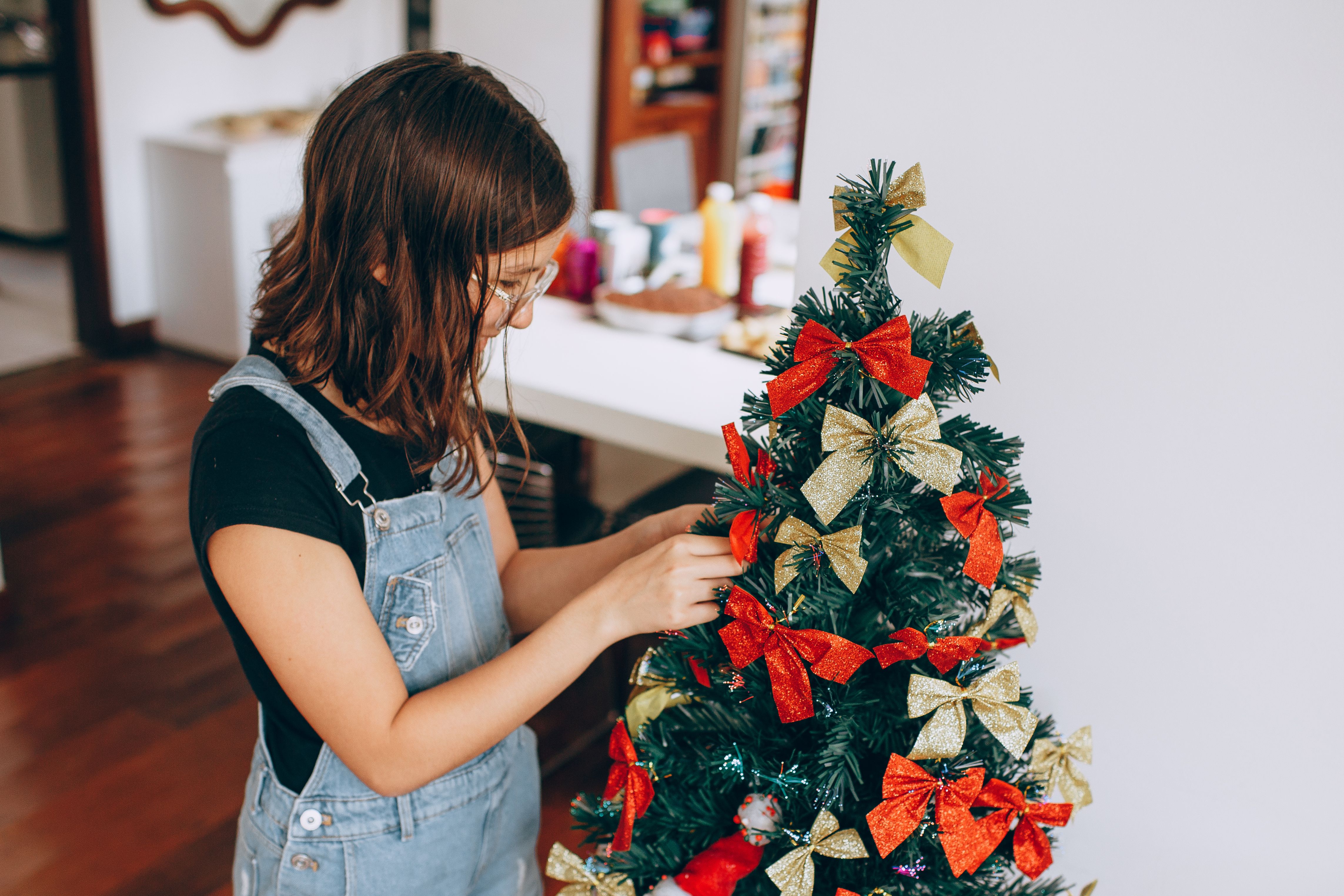 diy christmas decoration ideas at home A young girl in overalls decorates a small Christmas tree indoors with festive bows.