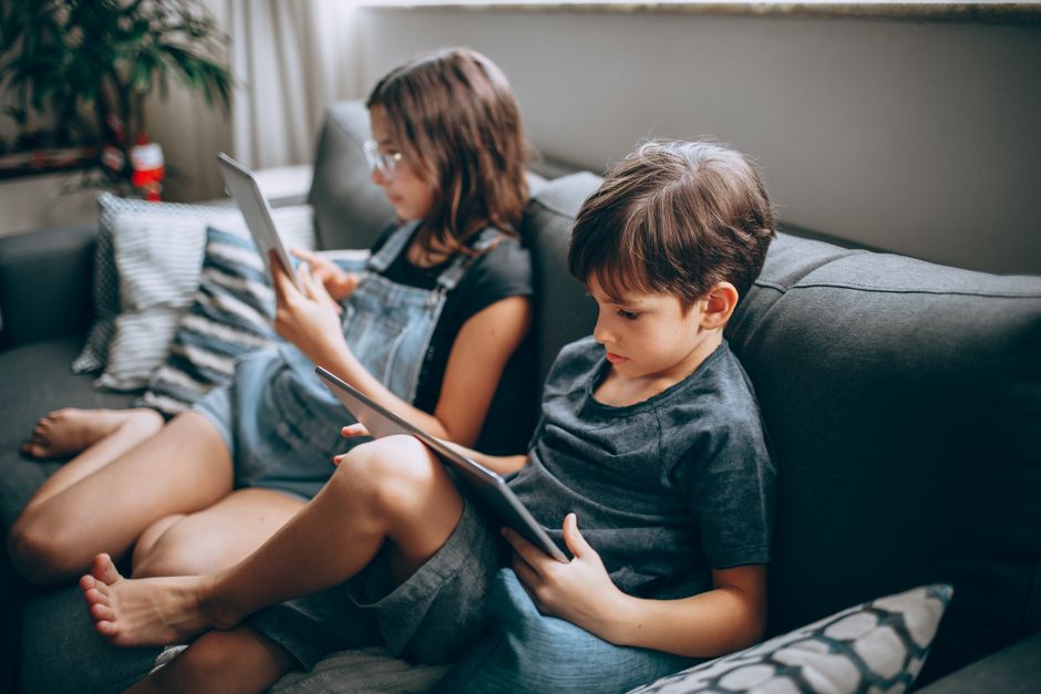 Two children sitting on a sofa using tablets indoors, focused on the screens.