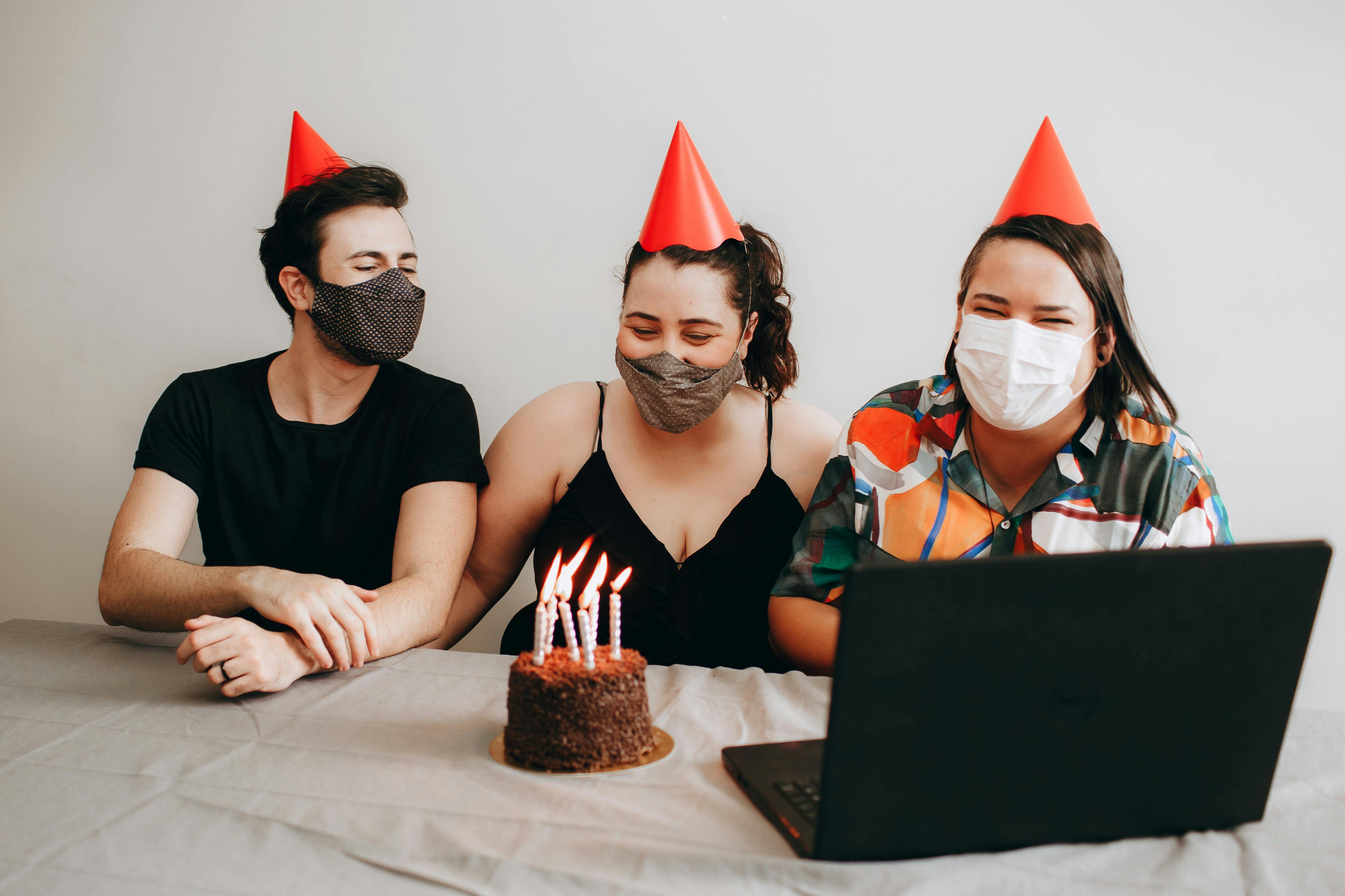 man and women in masks sitting with birthday cake and laptop