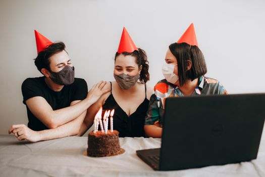 Three friends celebrating a birthday virtually with masks, party hats, and a cake.