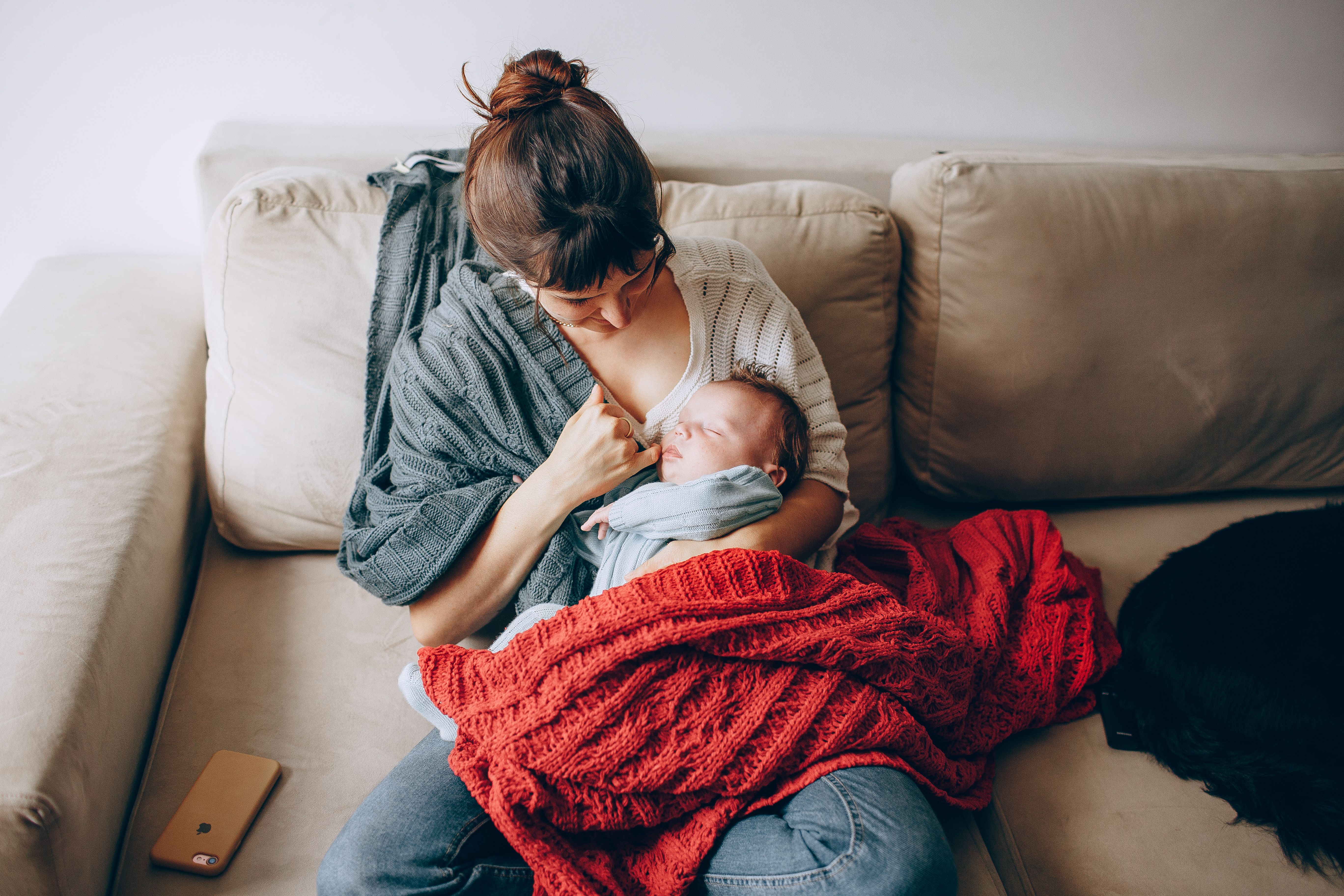A mother holding her baby on a couch, wrapped in warm blankets at home