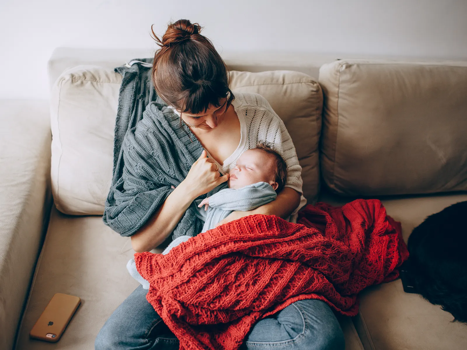 A mother holding her baby on a couch, wrapped in warm blankets at home