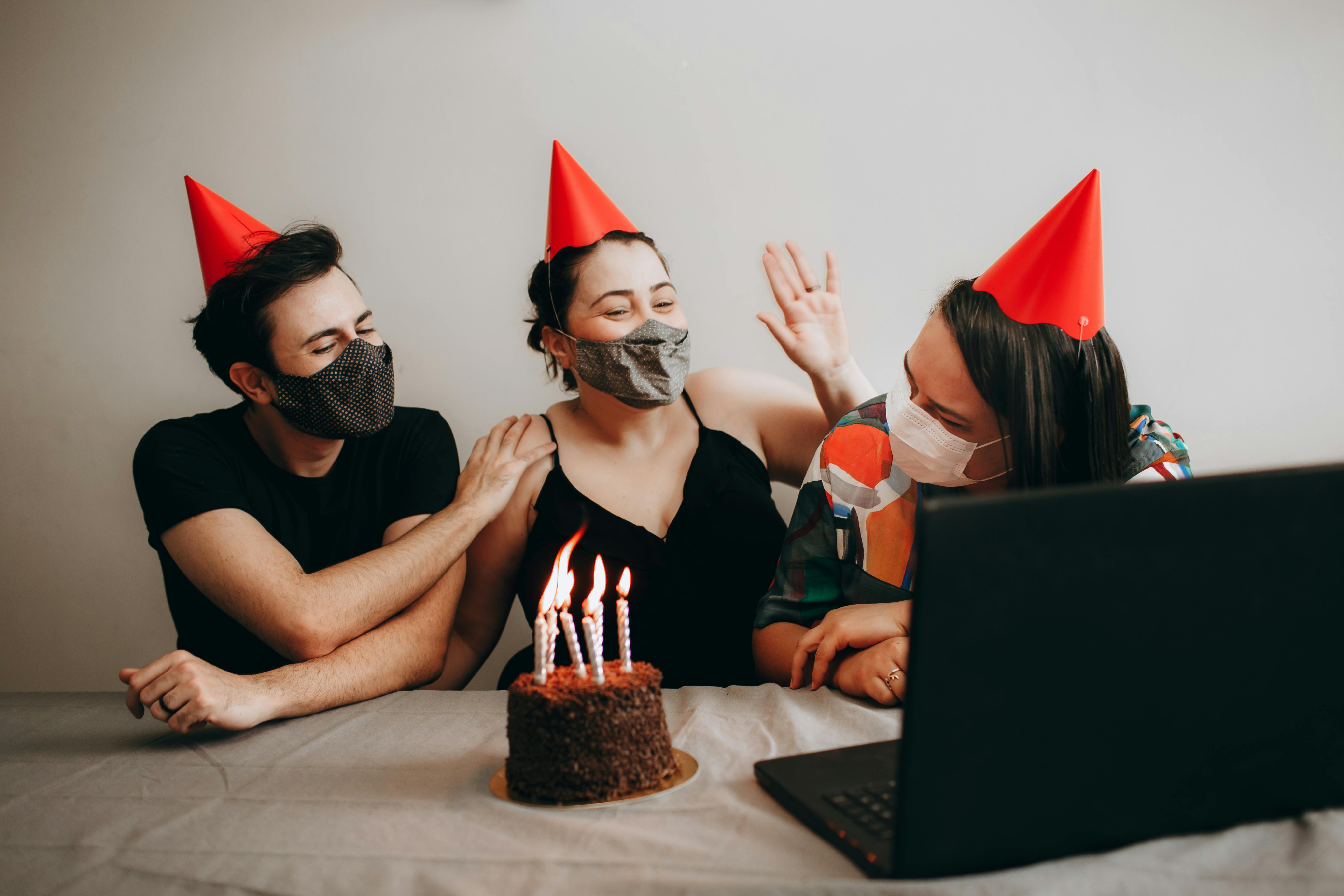 women and man in party hats sitting with birthday cake and laptop