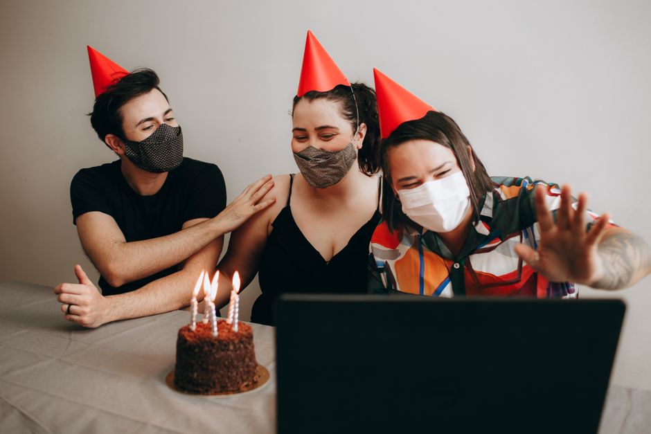 Three friends celebrate a birthday virtually wearing masks and party hats with a cake.