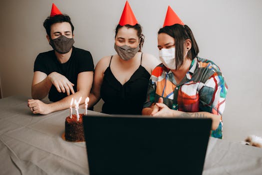 Three people celebrate a birthday with masks, party hats, and a cake via video call indoors.