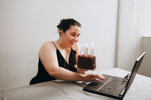Woman smiling and holding birthday cake with candles during a virtual celebration via laptop.
