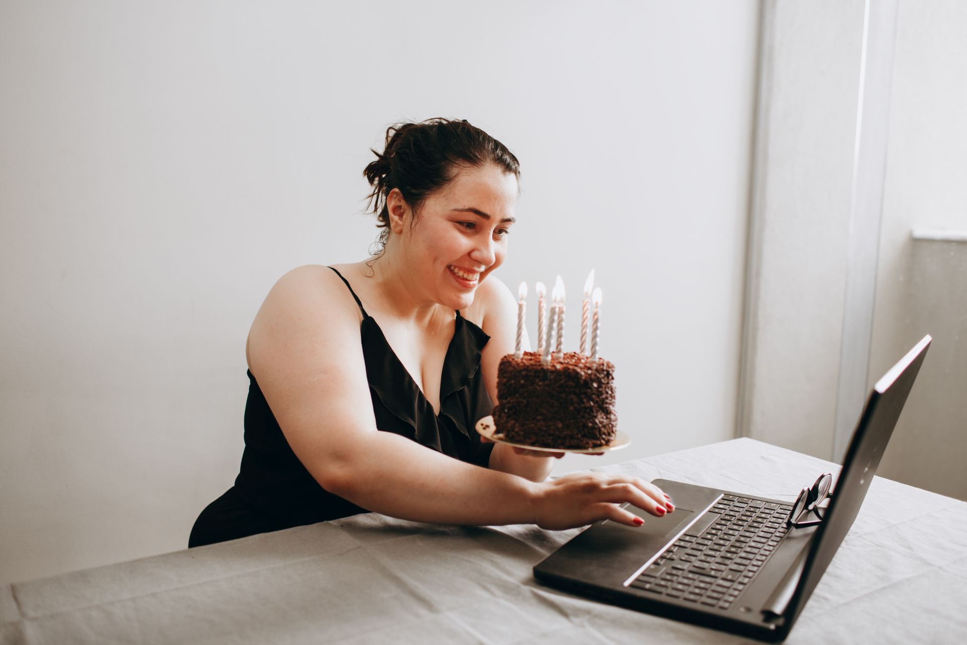 Woman smiling and holding birthday cake with candles during a virtual celebration via laptop.