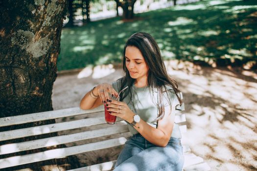 A young woman enjoys a refreshing drink while sitting on a park bench on a sunny day.