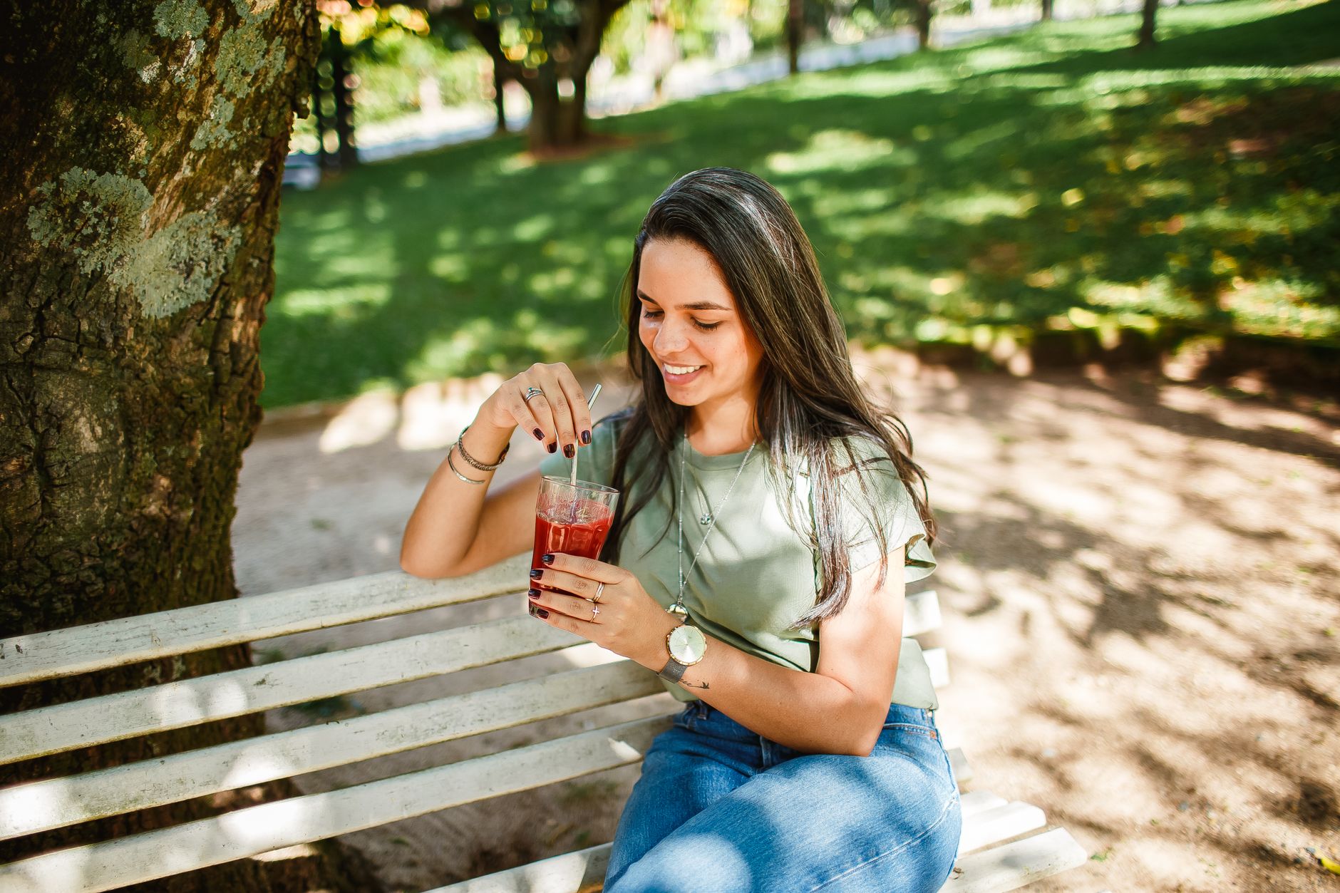 Smiling woman sitting on a bench enjoying a cold beverage in a sunny park.
