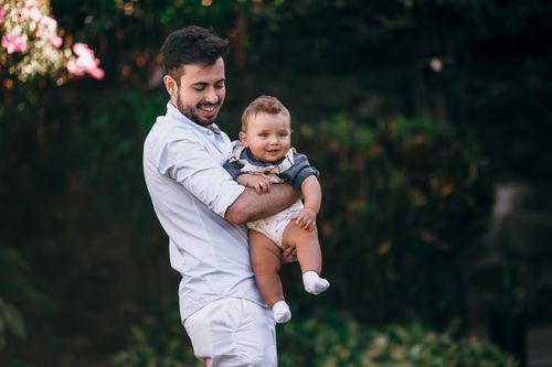Free Father joyfully holds his baby in a sunny park setting, capturing a moment of warm affection. Stock Photo