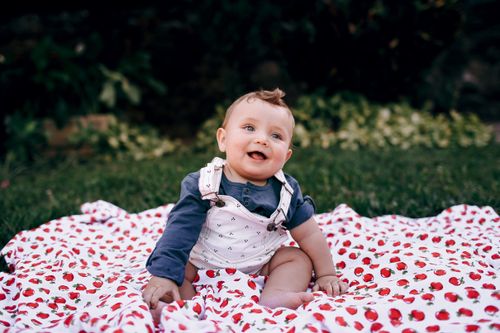 Free A happy baby sitting on a cherry-printed blanket outdoors, enjoying a sunny day. Stock Photo