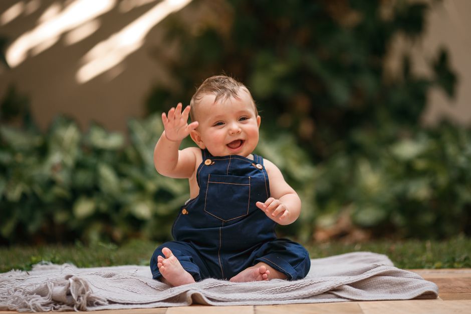 Adorable baby in denim overalls waving and smiling while sitting outdoors.