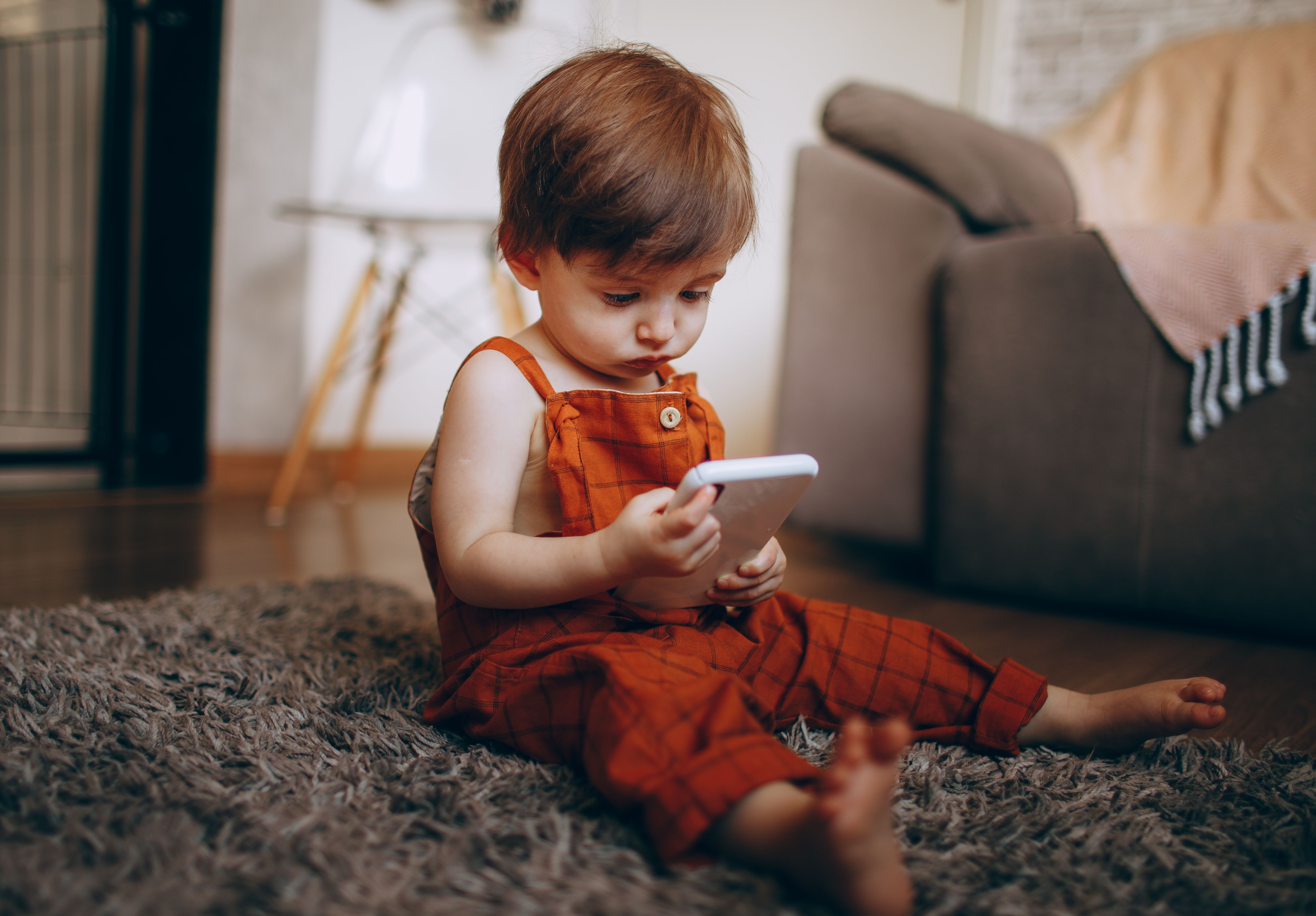 A cute toddler in orange overalls playing with a smartphone on a cozy carpet indoors.