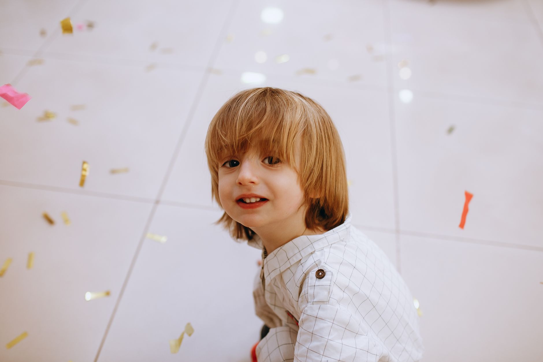 Young boy with long hair in a playful moment surrounded by festive confetti indoors.