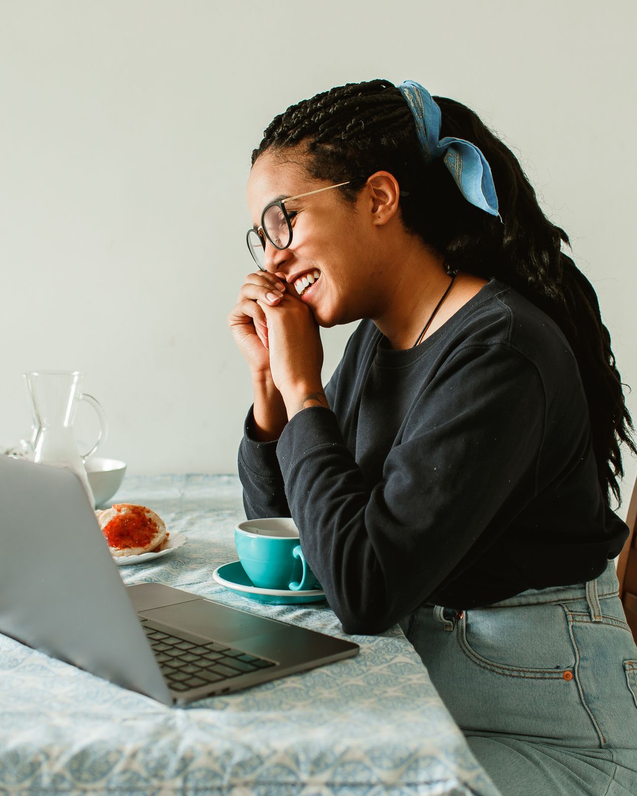 Retrato de uma mulher de óculos e cabelo preso, sorrindo diante do notebook em um café.