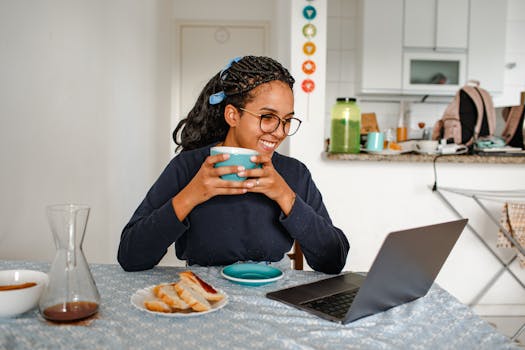 A woman smiles while drinking from a cup near her laptop, enjoying breakfast in a cozy home setting.