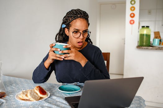 Young woman with glasses looks surprised while drinking coffee and using a laptop indoors.