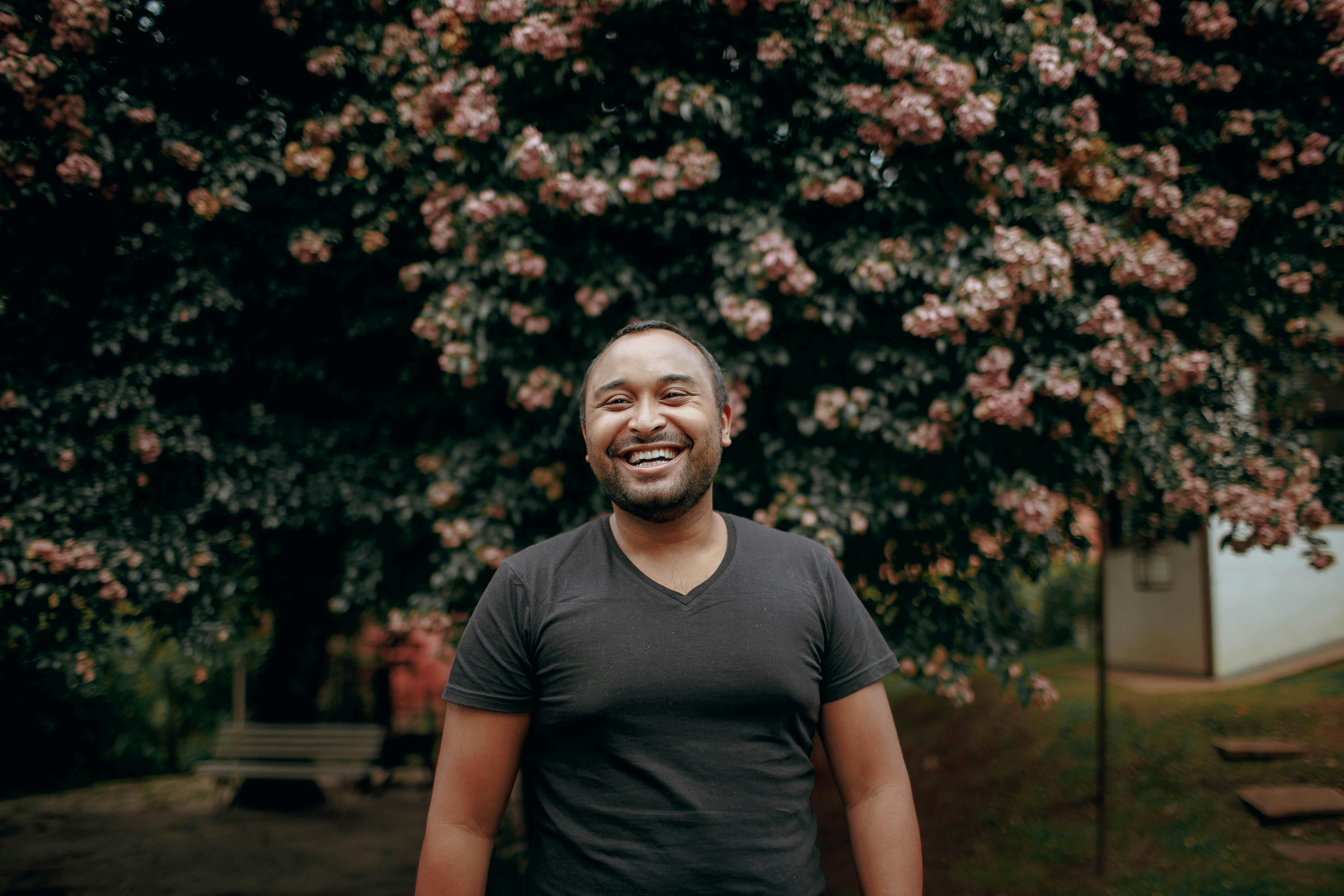 A man in a black shirt smiles in front of a tree