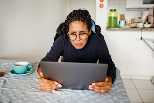 A woman wearing glasses looks stressed while using her laptop in a cozy kitchen setting.