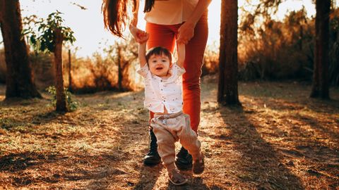 Mother helping her baby walk in a sunlit park, capturing a joyful moment.