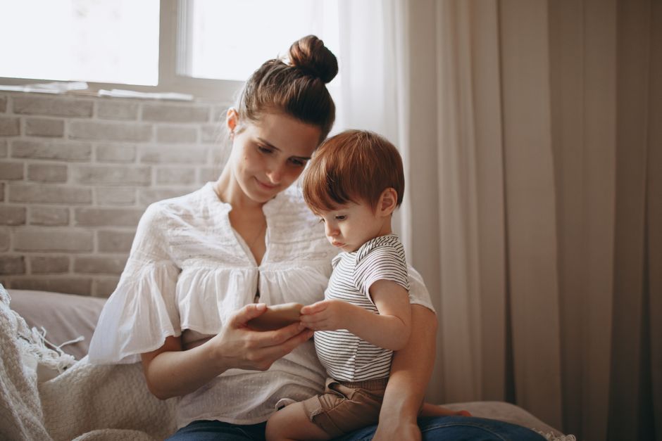 A mother and her young child enjoy a moment together indoors, sharing a smartphone.