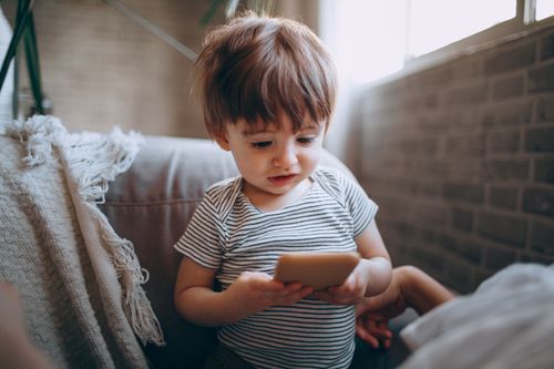 Free A cute toddler in a striped shirt focuses on a smartphone indoors, near a window. Stock Photo