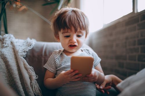 Free A toddler in a striped shirt explores a smartphone while sitting indoors on a cozy couch. Stock Photo