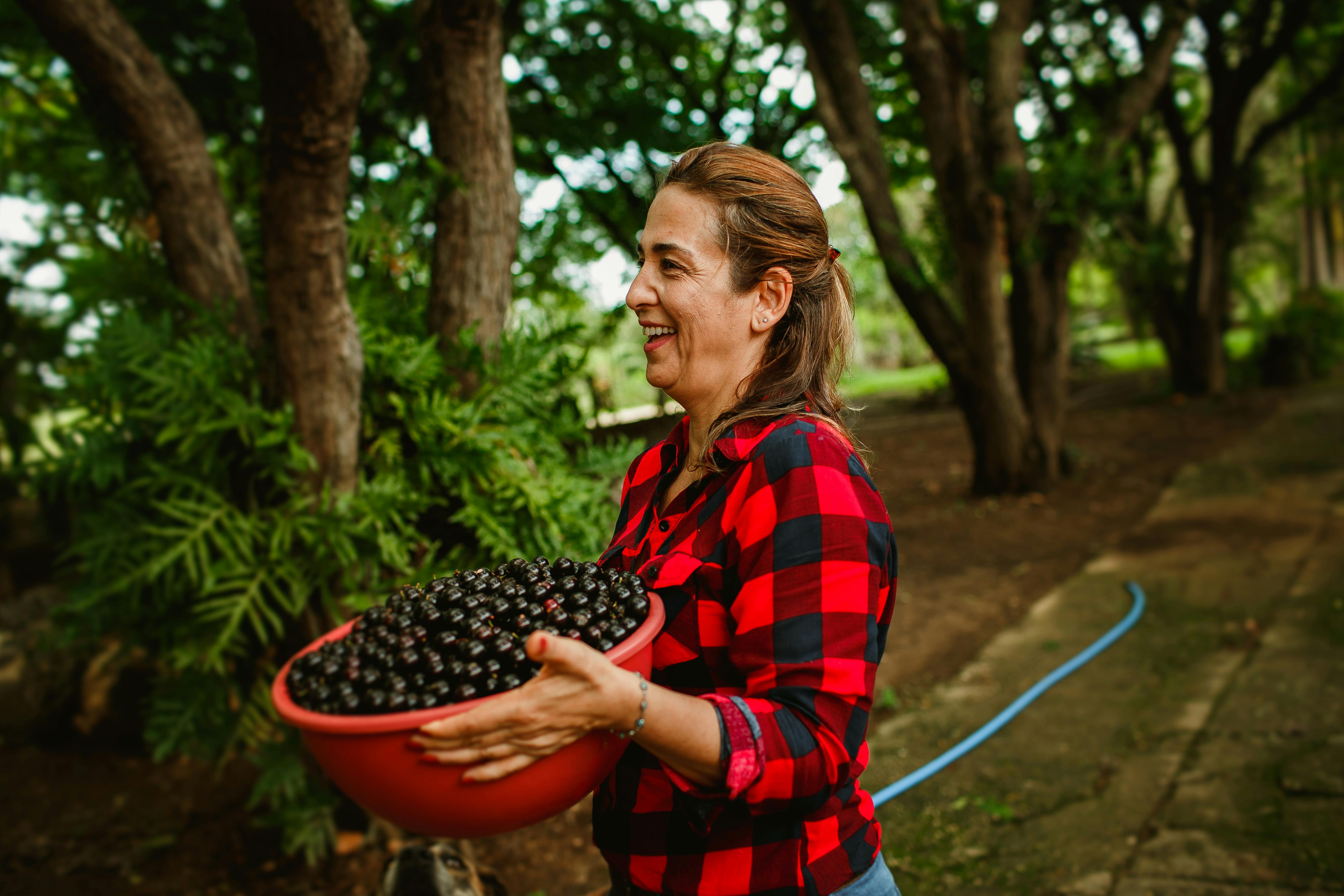 A happy woman in a plaid shirt holds a bowl of fresh blackberries in a lush green park.