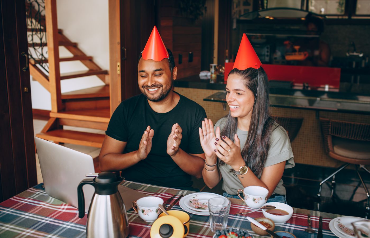 Two people clapping and smiling during a virtual celebration, wearing party hats and seated at a table.