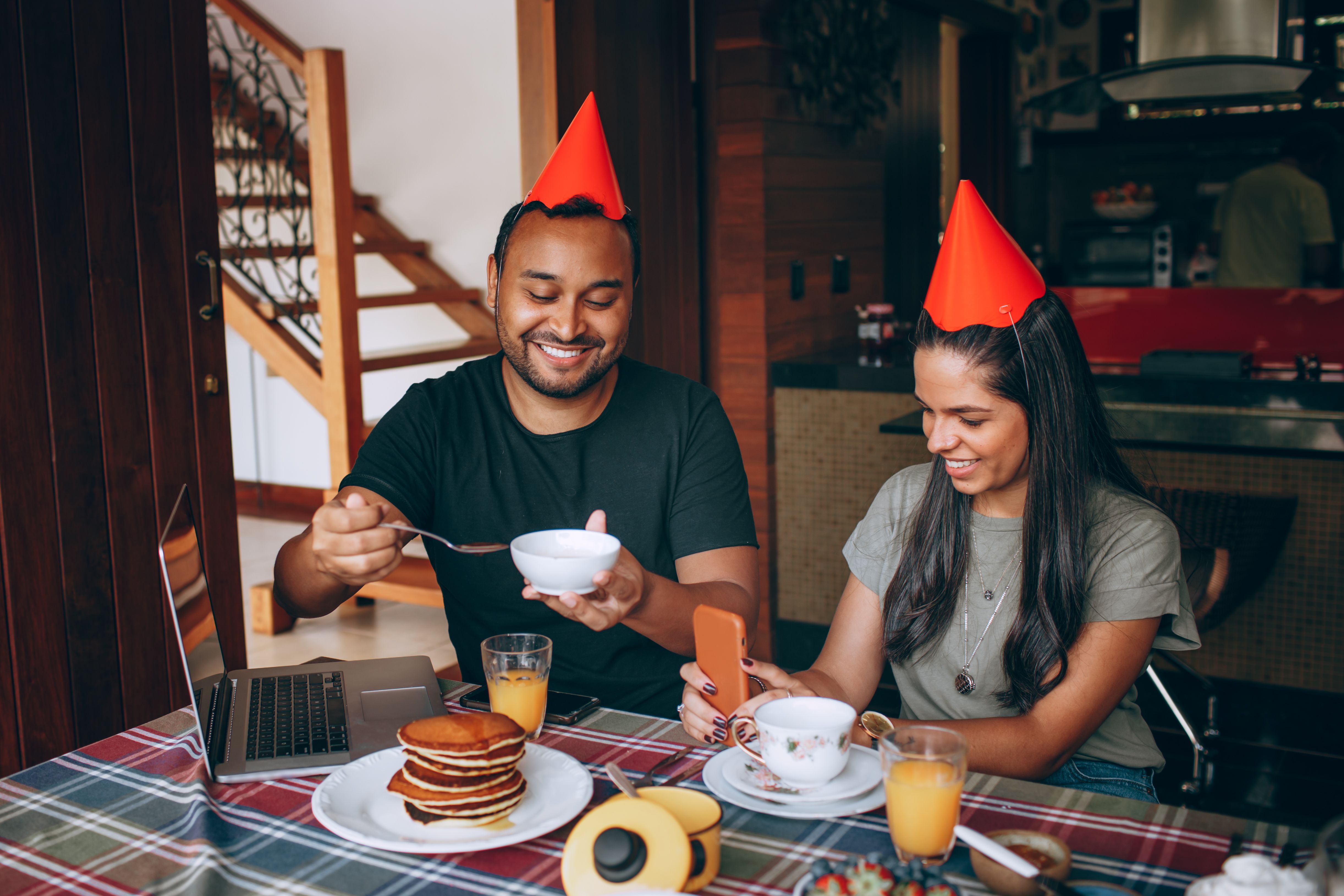 A couple celebrating a birthday breakfast indoors with pancakes, orange juice, and party hats.