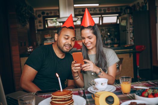 Two friends enjoying a birthday brunch indoors, wearing party hats and using a smartphone.