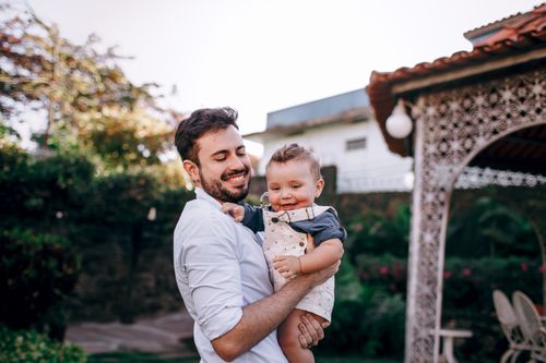 Free A joyful father holding his baby in a summer garden, surrounded by lush greenery. Stock Photo